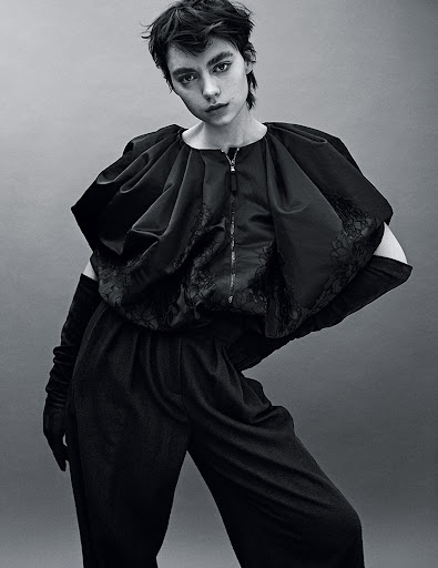 Monochrome portrait of a model wearing a dark, structured outfit with elbow-length gloves, posing in front of a grey backdrop.