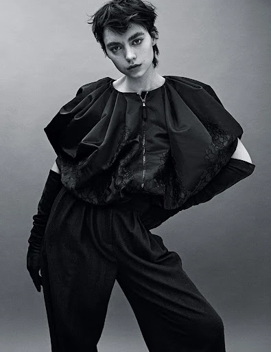 Monochrome portrait of a model wearing a dark, structured outfit with elbow-length gloves, posing in front of a grey backdrop.
