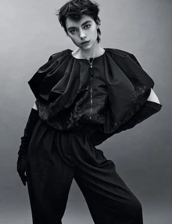 Monochrome portrait of a model wearing a dark, structured outfit with elbow-length gloves, posing in front of a grey backdrop.