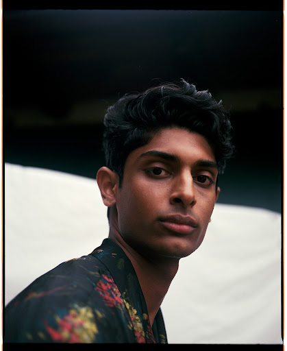 Portrait of a man with dark curly hair wearing a dark floral patterned shirt against a neutral background.