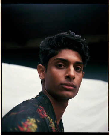 Portrait of a man with dark curly hair wearing a dark floral patterned shirt against a neutral background.