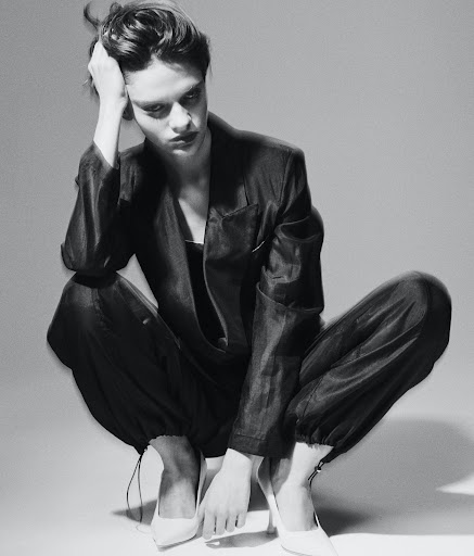 Adult female model in a dark pantsuit and white high heels is squatting in a photo studio with a neutral background, gazing down.