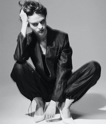 Adult female model in a dark pantsuit and white high heels is squatting in a photo studio with a neutral background, gazing down.