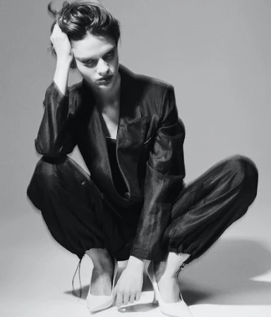 Adult female model in a dark pantsuit and white high heels is squatting in a photo studio with a neutral background, gazing down.