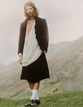 A person standing in a mountain landscape wearing a black blazer, white top and skirt, white face makeup, white socks, and black shoes.
