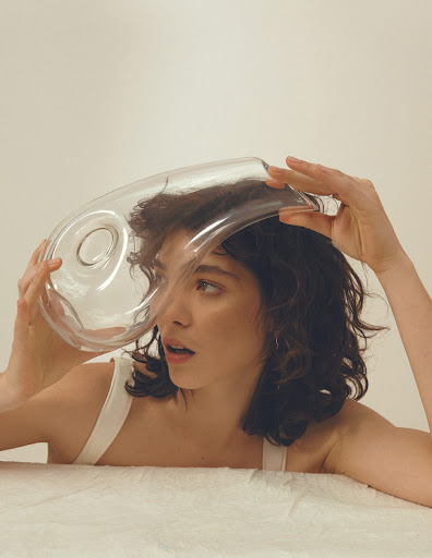A woman with shoulder-length dark hair and fair skin holds a glass vessel over her head in a studio, against a neutral background.