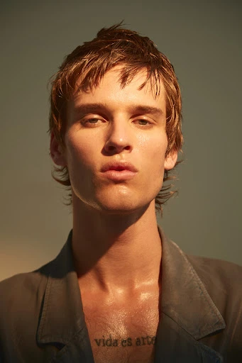 Close-up portrait of a young adult with light brown hair and a neutral facial expression, wearing a grey jacket and chest tattoo.