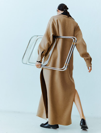 Fashion shot of a woman in a tan coat carrying a silver folding chair in a white studio, highlighting style and minimalist design.