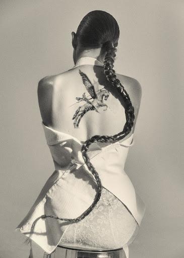 Back view of a woman with a long braid and Pegasus tattoo, wearing a white jacket and sitting on a chrome stool against a grey backdrop.