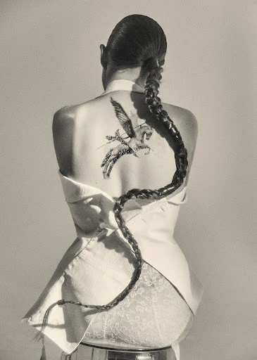 Back view of a woman with a long braid and Pegasus tattoo, wearing a white jacket and sitting on a chrome stool against a grey backdrop.