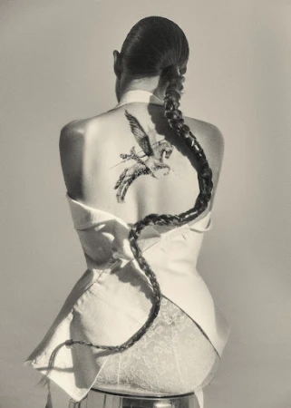 Back view of a woman with a long braid and Pegasus tattoo, wearing a white jacket and sitting on a chrome stool against a grey backdrop.