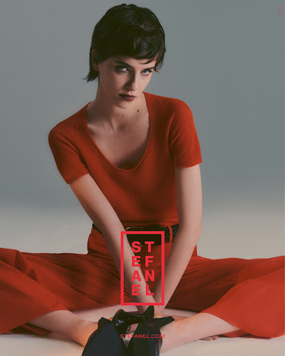Fashion image of a young woman in a red knit outfit and black high heels sitting on the floor looking at the camera in a photo studio.