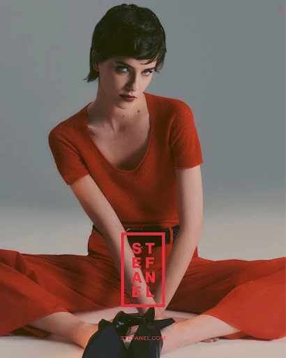 Fashion image of a young woman in a red knit outfit and black high heels sitting on the floor looking at the camera in a photo studio.