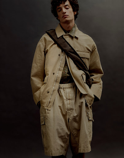 Young man poses in a tan jacket and khaki shorts, accented by a dark brown cross-body bag, set against a muted gray backdrop.