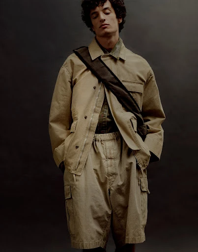 Young man poses in a tan jacket and khaki shorts, accented by a dark brown cross-body bag, set against a muted gray backdrop.