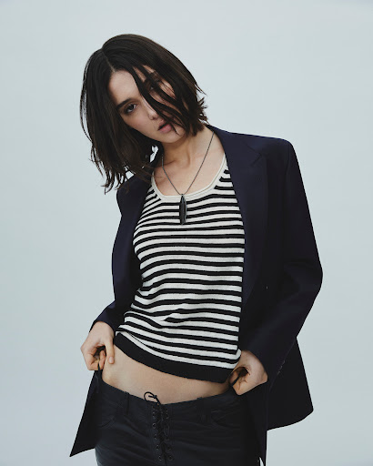 A woman with short dark hair poses in a striped tank top, black jacket, and dark pants against a plain background in a studio shot.