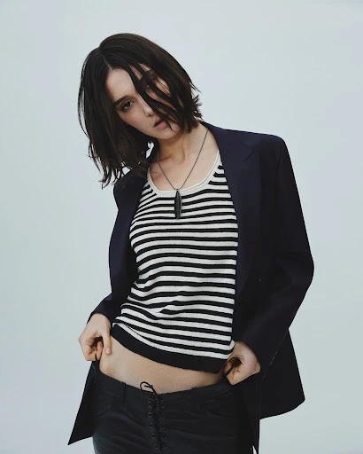 A woman with short dark hair poses in a striped tank top, black jacket, and dark pants against a plain background in a studio shot.