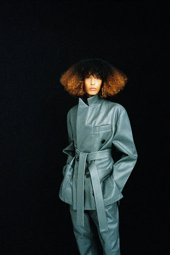 Woman in a grey belted jacket and matching pants poses against a black background in a fashion shot, showcasing textures and silhouette.