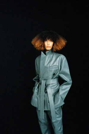 Model with voluminous two-toned hair wearing a grey leather belted jacket and matching trousers against a black background