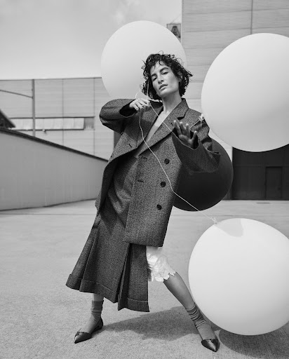 Black and white portrait of woman in oversized coat holding three balloons outdoors on a concrete surface with a building in the background.