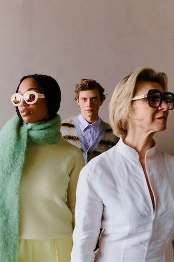 Three models with sunglasses and varied knitwear and shirts posed against a neutral wall