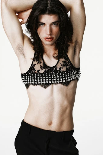 Adult male with long dark hair poses in black lace and studded bra and black trousers against a white backdrop in a studio shot.