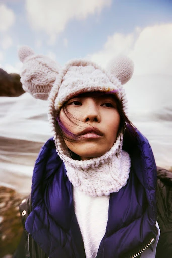 Portrait of a stylish adult woman wearing a knitted hat with ear flaps and puffer jacket with water and cloudy sky in the background.