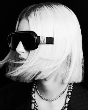 Close-up portrait of a model with sleek platinum blonde hair and dark sunglasses against a black background, styled by Simone Prusso.
