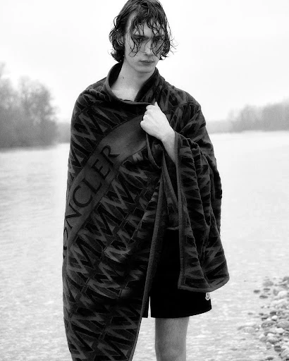 Black and white portrait of a young adult male wrapped in a towel by a river, facing the camera with a serious expression.