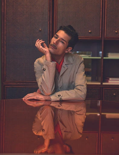 Portrait of an adult male with a mustache wearing a blazer sitting at a reflective table in a dimly lit room with a bookshelf.
