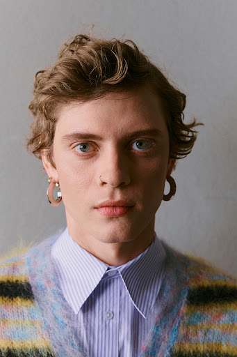 Close-up portrait of a young adult man with wavy light brown hair, wearing a striped cardigan and collared shirt against a grey backdrop.