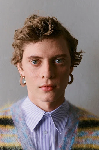 Close-up portrait of a young adult man with wavy light brown hair, wearing a striped cardigan and collared shirt against a grey backdrop.