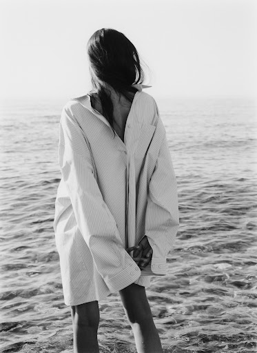 Monochrome portrait of a woman standing in water facing away, wearing an oversized striped button-down shirt.