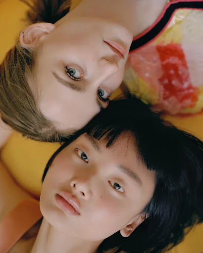 Two young women lie head-to-head against a yellow backdrop. A blonde woman with blue eyes wears a colorful top; an Asian woman has dark hair.