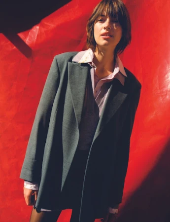 Woman with layered dark hair wearing a grey oversized blazer over a pink shirt against a textured red background.