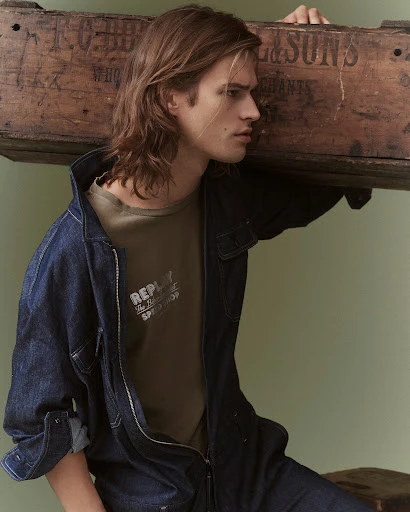 Portrait of a fair-skinned male model with long brown hair wearing a denim jumpsuit and olive T-shirt posing indoors with a wooden crate.