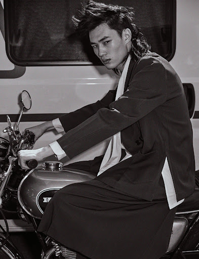 Monochrome portrait of a stylish adult male with a mullet, seated on a motorcycle in front of a white vehicle, with a serious expression.