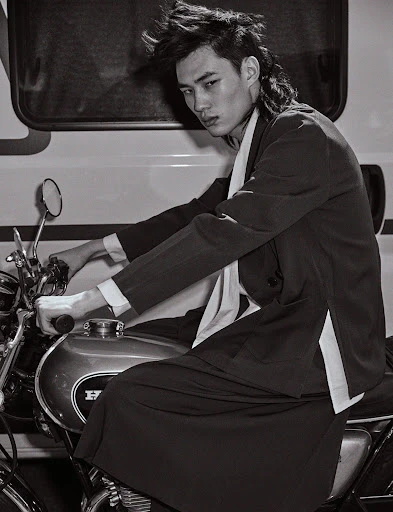 Monochrome portrait of a stylish adult male with a mullet, seated on a motorcycle in front of a white vehicle, with a serious expression.