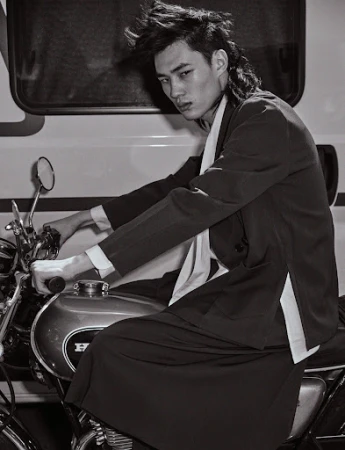 Monochrome portrait of a stylish adult male with a mullet, seated on a motorcycle in front of a white vehicle, with a serious expression.
