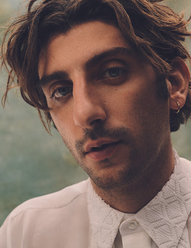 Portrait of a young adult male with dark hair and blue eyes, wearing a white shirt with a textured collar against a soft background.