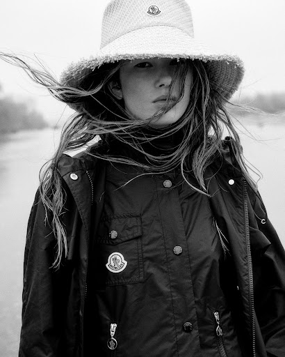 Model wearing a bucket hat and dark jacket outdoors in a black and white portrait, hair blowing in the wind.