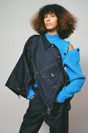 Model with voluminous curly hair wearing a blue knit sweater and dark denim jacket against a plain background