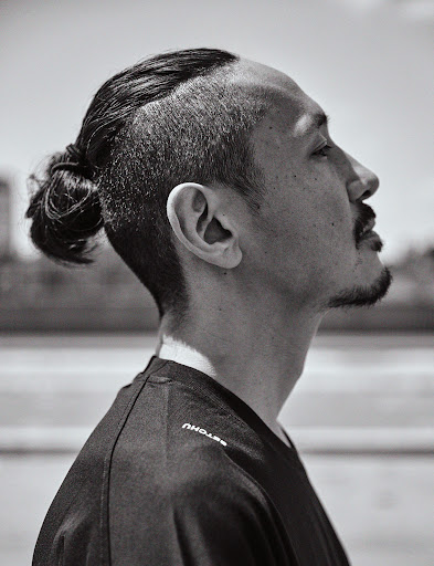 Black and white profile portrait of an adult male with an undercut and hair bun, wearing a black shirt, in an outdoor setting.