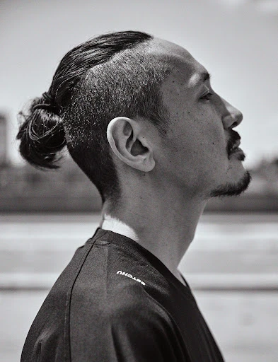 Black and white profile portrait of an adult male with an undercut and hair bun, wearing a black shirt, in an outdoor setting.