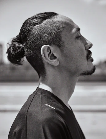 Black and white profile portrait of an adult male with an undercut and hair bun, wearing a black shirt, in an outdoor setting.