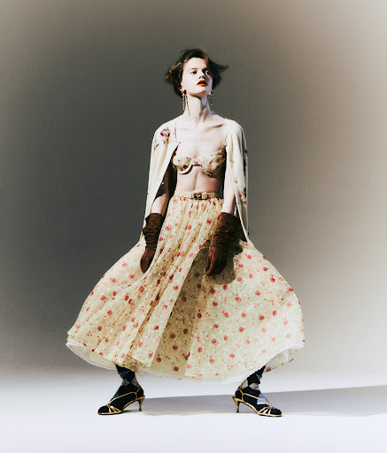 A woman stands in a light grey studio wearing a floral bra top, a matching skirt and cardigan, with high heels and dark tights.