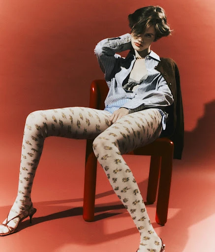 A fashionable woman sitting on a red chair in a photo studio, wearing a blue and white shirt, decorative patterned tights, and heeled sandals.
