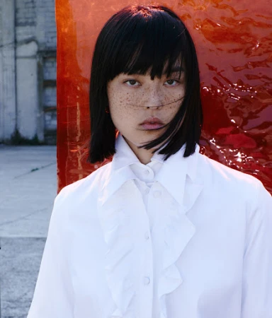 Portrait of a woman with dark bob hair and freckles wearing a white ruffled shirt against an orange panel and outdoor background
