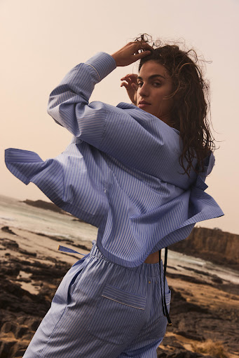 A brunette woman in a blue-striped shirt and pants poses on a rocky outcrop, looking toward the camera on an overcast day.