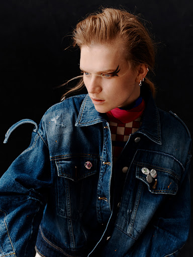 Portrait of a young adult woman with bold winged eyeliner, wearing a distressed denim jacket, against a dark background in studio lighting.